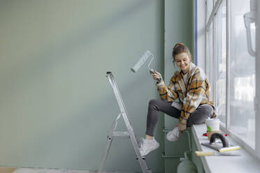 Smiling woman with paint roller sitting on window sill at home - VIVF00131