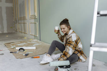 Smiling woman with paint roller having coffee in messy apartment - VIVF00140