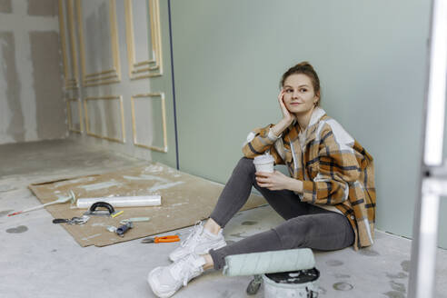 Nachdenkliche Frau mit Einweg-Kaffeebecher sitzt zu Hause auf dem Boden - VIVF00142