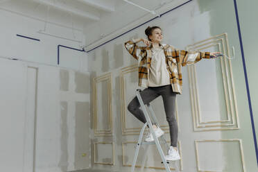 Smiling young woman dancing with paint roller on ladder in apartment - VIVF00143
