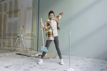 Cheerful woman in plaid shirt dancing with broom in apartment - VIVF00157