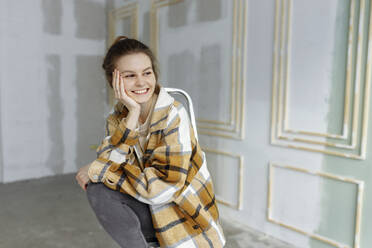 Smiling young woman in plaid shirt day dreaming with hand on chin in apartment - VIVF00170