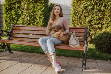Lächelnde Frau mit Hund auf einer Bank im Park sitzend - MDOF00173