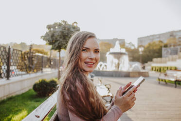 Happy woman holding smart phone on bench in park - MDOF00183