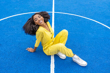 Glückliche junge Frau sitzt auf blauem Basketballplatz - DLTSF03396