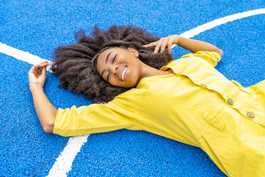 Glückliche Frau mit Afrofrisur auf blauem Basketballplatz liegend - DLTSF03401