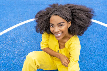 Smiling young woman sitting on blue basketball court - DLTSF03410