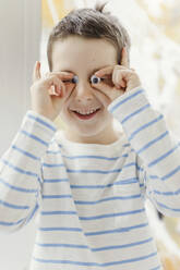 Happy boy holding small googly eyes over face at home - ONAF00245