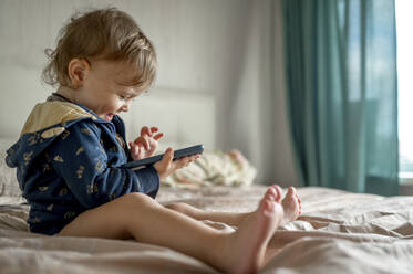 Glücklicher kleiner Junge, der ein Spiel auf seinem Smartphone im Schlafzimmer spielt - ANAF00480