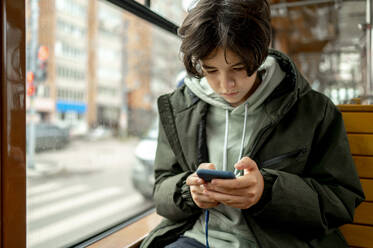Junge schaut auf sein Handy, sitzt am Fenster in der Straßenbahn - ANAF00484