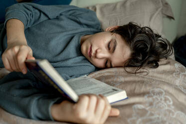 Boy reading novel lying on bed at home - ANAF00494