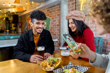 Junges, fröhliches, ethnisches Paar in Freizeitkleidung, das sich unterhält und liebevoll anschaut, während es mit einem kalten, frischen Bier in der Nähe eines Freundes am Tisch sitzt und sein Smartphone benutzt - ADSF40221