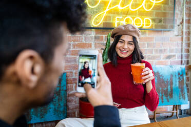 Mann mit Smartphone, der ein Bild von einer glücklich lächelnden ethnischen Freundin macht, die ein Glas eines leckeren Cocktails in einem modernen Restaurant hält - ADSF40223