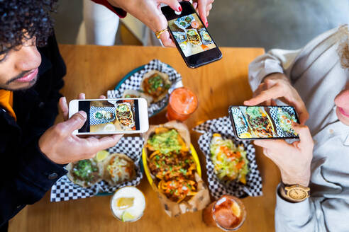 Top view crop anonymous friends taking pictures of delicious food on smartphones while gathering at table in restaurant - ADSF40224
