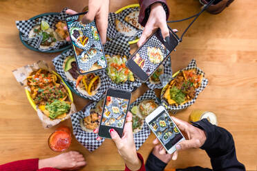 Top view crop anonymous friends taking pictures of delicious food on smartphones while gathering at table in restaurant - ADSF40227