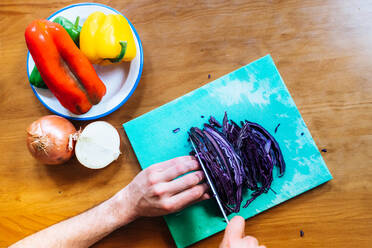 Draufsicht auf einen anonymen Koch, der Rotkohl auf einem Schneidebrett neben einem Teller mit reifen Paprika und Zwiebeln schneidet - ADSF40236