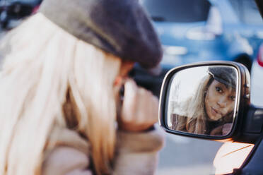 Young African American woman looking at wing mirror and applying mascara while standing on city street on sunny day - ADSF40264