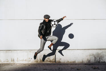 African American guy in trendy outfit jumping kicking football boy near grungy wall on city street - ADSF40268