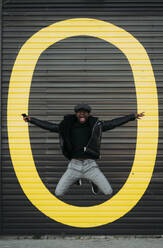 Junger schwarzer Mann springt auf der Straße vor einer Mauer mit der gelben Zahl Null - ADSF40280