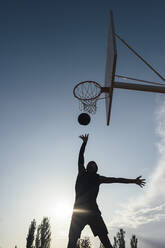 Kräftiger, energiegeladener afroamerikanischer Sportler, der sich auf dem Spielplatz an einen Basketballkorb hängt, nachdem er den Ball im Netz versenkt hat - ADSF40296