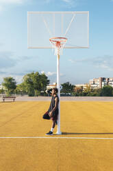 Müder afroamerikanischer Sportler in Uniform lehnt neben dem Korb auf einem gelb markierten Spielplatz mit Ball am hellen Tag - ADSF40297