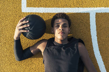 Draufsicht auf einen müden afroamerikanischen Sportler in Uniform, der sich auf einem gelb markierten Spielplatz mit einem Ball am hellen Tag ausruht - ADSF40298