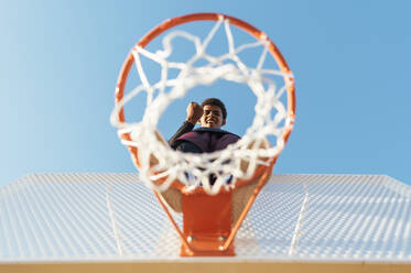 Blick von unten auf einen afroamerikanischen Sportler, der auf einem Basketballring steht und mit einer geballten Faust als Siegeszeichen in die Kamera schaut - ADSF40302