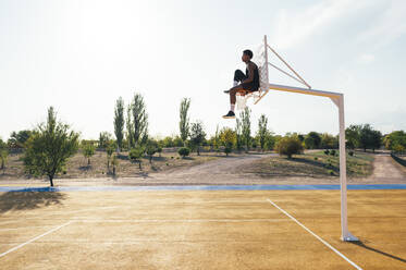 Seitenansicht eines afroamerikanischen Sportlers, der auf einem Basketballring sitzt und auf einen leeren Spielplatz blickt - ADSF40303