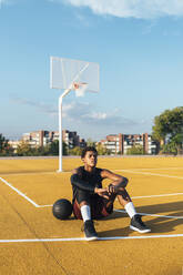 Tired African American sportsman in uniform having rest sitting on yellow marked playground with ball in bright day - ADSF40306