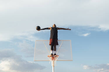 Ein afroamerikanischer Sportler, der auf dem Basketballring steht und zum Himmel schaut und die Hände zur Seite streckt - ADSF40307
