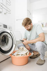 Ein Mann benutzt sein Smartphone an der Waschmaschine im Badezimmer zu Hause - EYAF02320