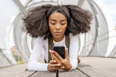 Junge Frau mit lockigem Haar, die auf einer Fußgängerbrücke ihr Smartphone benutzt - DLTSF03439