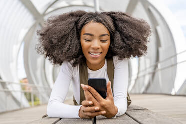 Smiling woman looking at smart phone on footbridge - DLTSF03440