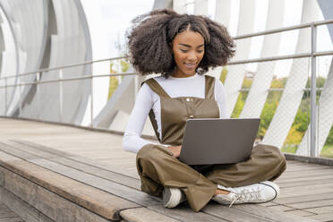 Woman using laptop sitting cross-legged on footbridge - DLTSF03443