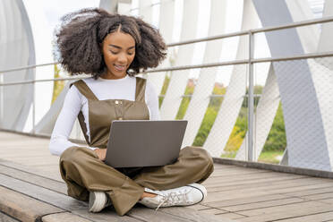 Young woman using laptop sitting cross-legged on footbridge - DLTSF03444
