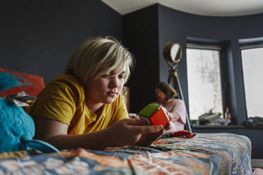 Junge spielt mit Puzzle-Würfel auf dem Bett liegend zu Hause - EYAF02322