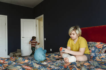 Junge mit Gipsverband am Bein, der mit einem Puzzle-Würfel spielt, der zu Hause auf dem Bett liegt - EYAF02325