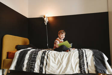 Glücklicher Junge mit Buch auf dem Bett sitzend - EYAF02329