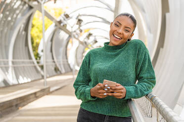 Happy young woman holding mobile phone on bridge - DLTSF03449