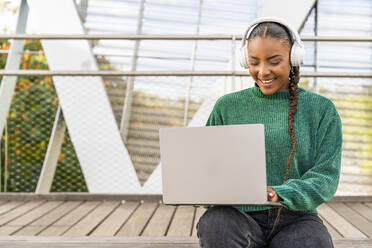 Glückliche Frau, die über drahtlose Kopfhörer Musik hört und einen Laptop auf einer Brücke benutzt - DLTSF03450