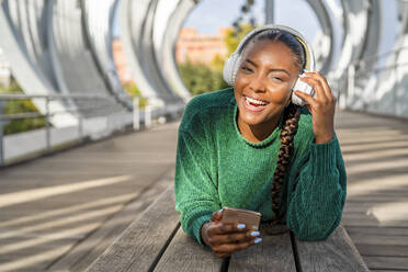Glückliche Frau hört Musik über Kopfhörer und hält ihr Handy auf einer Brücke liegend - DLTSF03468