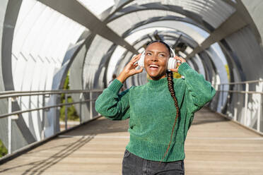 Happy young woman enjoying music through wireless headphones on bridge - DLTSF03469