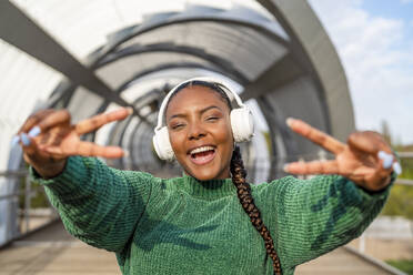 Glückliche junge Frau, die über drahtlose Kopfhörer Musik hört und auf der Brücke gestikuliert - DLTSF03470