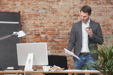 Businessman looking at document talking on speaker phone at work place - DHEF00664