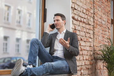 Geschäftsmann, der mit seinem Smartphone auf der Fensterbank sitzt - DHEF00667