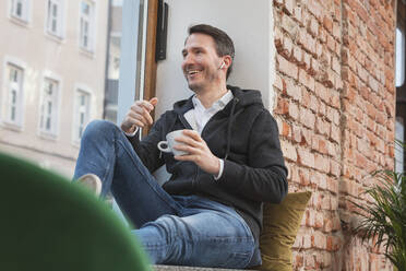 Glücklicher Geschäftsmann mit In-Ear-Kopfhörern und Kaffeetasse auf dem Fensterbrett sitzend - DHEF00669