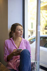 Lächelnde Geschäftsfrau mit Smartphone schaut durch das Fenster eines Cafés - JOSEF14723