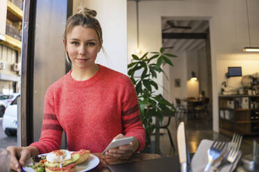 Smiling young freelancer sitting with mobile phone and plate of food at cafe - JOSEF14732