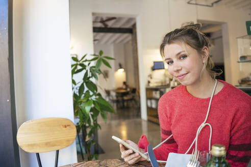 Lächelnder junger Freiberufler mit Smartphone im Café sitzend - JOSEF14748
