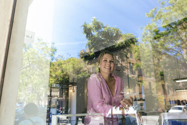 Glückliche Geschäftsfrau mit Smartphone durch Glas im Café gesehen - JOSEF14763